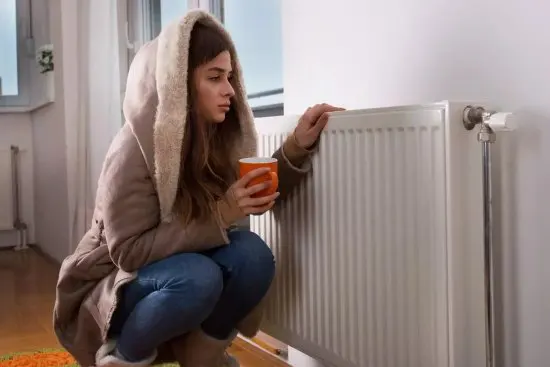 Mädchen im Mantel lehnt an einem Heizkörper wegen eines schlecht funktionierenden Kessels durch Kalkablagerungen.